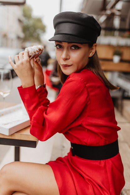 Divertido retrato de linda chica hambrienta en café. Joven morena come croissant con gusto