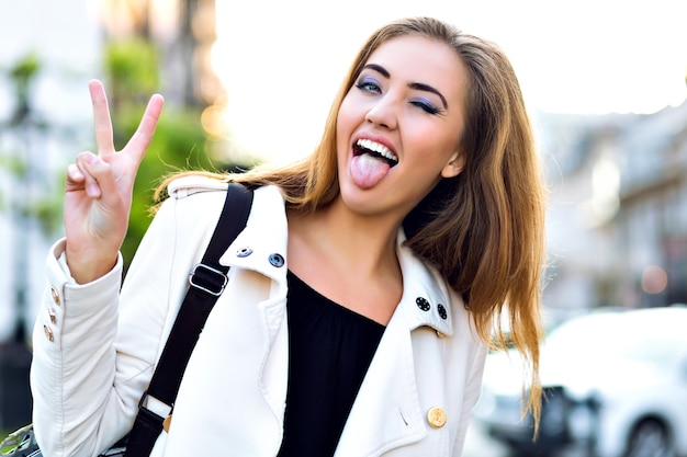 Divertido retrato de niña rubia feliz, haciendo muecas y mostrando la lengua en la calle, tiempo de otoño, descanso en la ciudad.