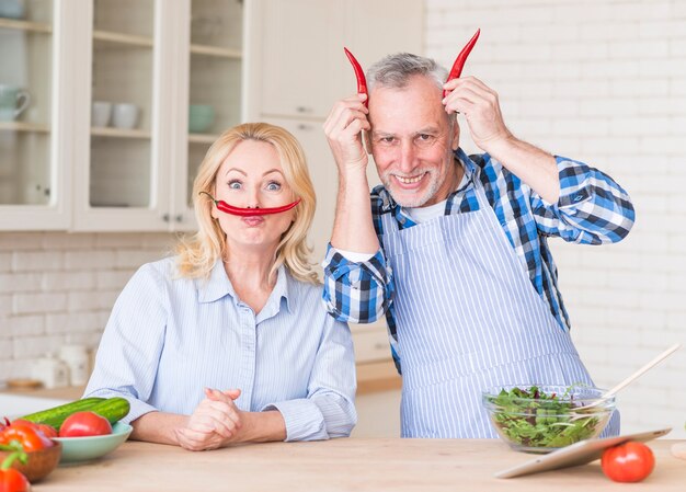 Divertido retrato de una pareja senior con chiles rojos en la cocina