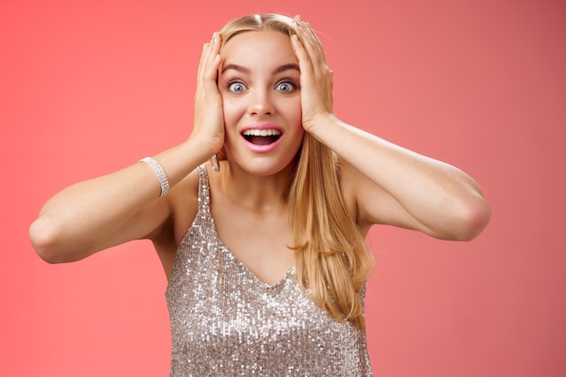 Divertido sorprendido emocionado joven mujer atractiva rubia en vestido plateado brillante con estilo mantener las manos en la cabeza abrir los ojos sin palabras mirar cámara increíble emoción fan ver persona famosa, fondo rojo.