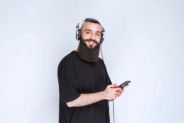 Dj con barba usando audífonos y poniendo música desde su playlist en su smartphone.