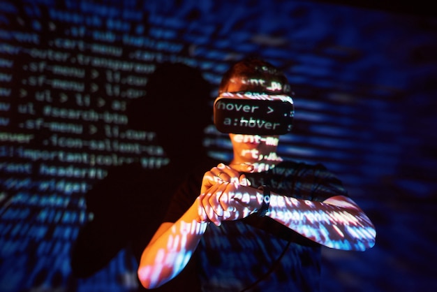 La doble exposición de un hombre caucásico y un casco de realidad virtual VR es presumiblemente un jugador o un pirata informático que descifra el código en una red o servidor seguro, con líneas de código.