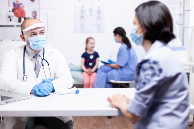 Docto conversando con los padres mientras la enfermera consulta al niño con máscara protectora. Médico especialista que brinda servicios de atención médica consultas tratamiento en equipo de protección.