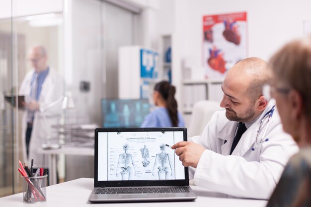 Doctor apuntando al esqueleto humano en la computadora portátil en la oficina del hospital durante la consulta de la anciana antes de la cirugía. Médico senior vistiendo bata blanca tomando notas en el portapapeles en el pasillo de la clínica.