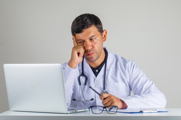 Doctor en bata blanca, estetoscopio sentado y mirando portátil y mirando con cuidado