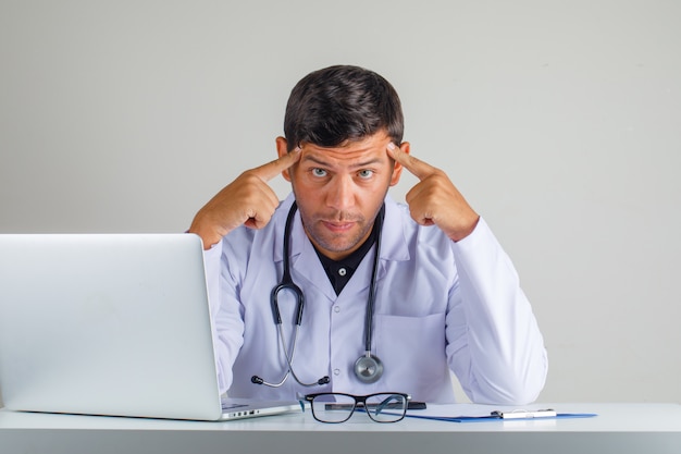 Foto gratuita doctor en bata blanca, estetoscopio tocando las sienes con los dedos y con aspecto cansado