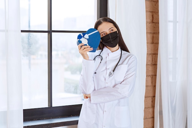 Doctor con estetoscopio y máscara negra sosteniendo una caja de regalo con forma de corazón azul y parece confundido e inseguro.