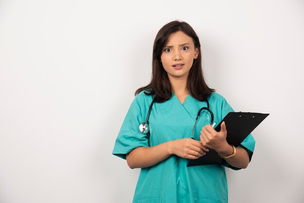 Doctor con estetoscopio y portapapeles parece sorprendido sobre fondo blanco. Foto de alta calidad