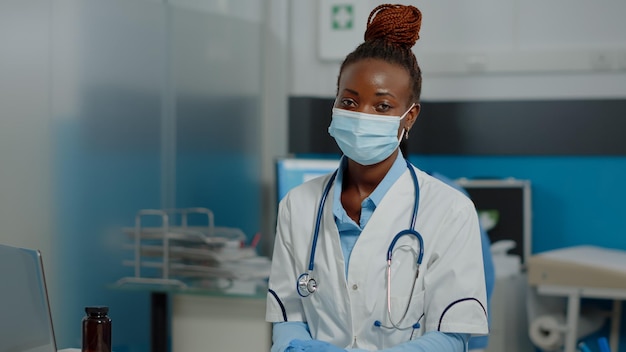 Doctor de etnia afroamericana sentado en el escritorio con laptop y documentos mientras usa mascarilla y bata blanca mirando a la cámara. mujer negra que trabaja como médico en el gabinete de salud