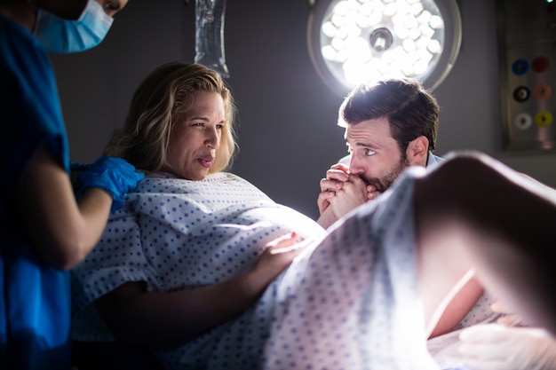 Doctor examinando a la mujer embarazada durante el parto mientras el hombre sosteniendo su mano en la sala de operaciones