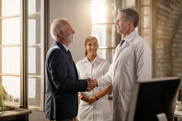 Doctor feliz dándose la mano con un hombre de negocios senior mientras la enfermera está de pie en el fondo