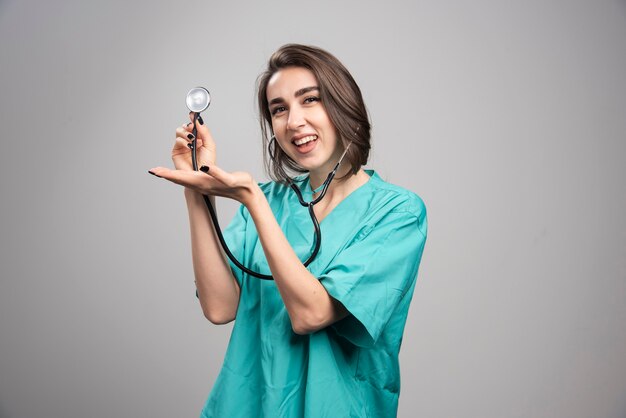 Doctor feliz que muestra el estetoscopio sobre fondo gris. Foto de alta calidad