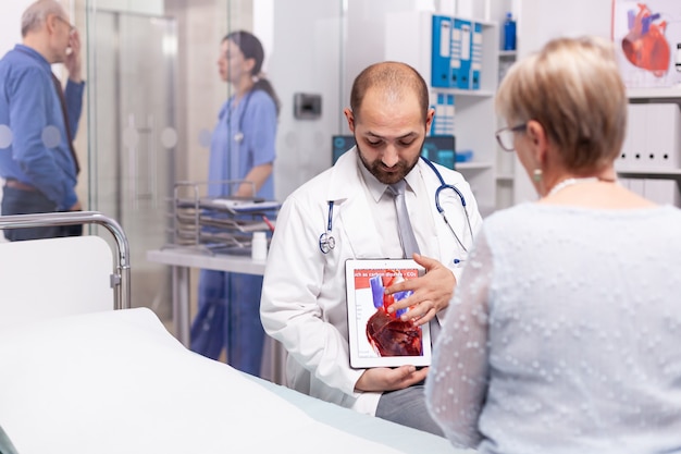 Doctor hablando sobre el efecto del ataque cardíaco apuntando a la tablet pc durante el examen de la mujer mayor