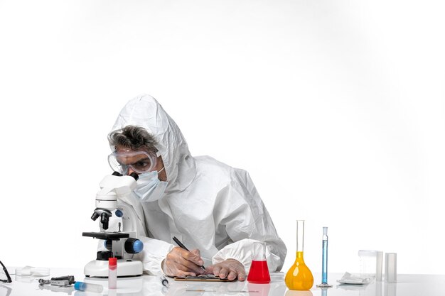 Doctor hombre en traje de protección escribiendo notas y usando microscopio en blanco