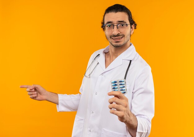 Doctor joven complacido con gafas médicas vistiendo bata médica con estetoscopio sosteniendo pastillas y apunta a un lado