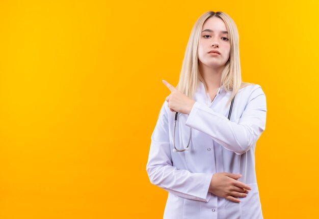 Doctor joven con estetoscopio en bata médica apunta al lado de la pared amarilla aislada