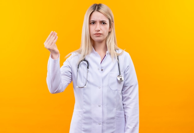 Doctor joven con estetoscopio en bata médica mostrando gesto de efectivo en pared amarilla aislada