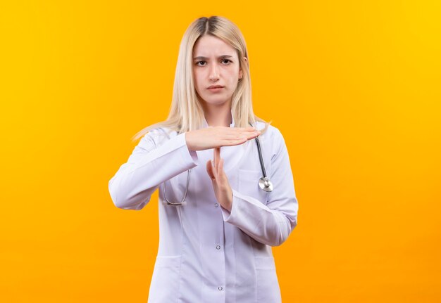 Doctor joven con estetoscopio en bata médica mostrando gesto de tiempo de espera en la pared amarilla aislada