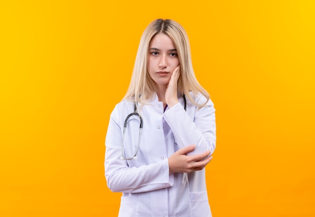 Doctor joven con estetoscopio en bata médica puso su mano en la mejilla en la pared amarilla aislada