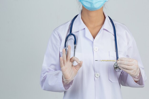 El doctor joven hermoso lleva la máscara mientras que sostiene el termómetro con los guantes de goma en la pared gris.