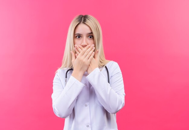 Doctor joven rubia con estetoscopio en bata médica boca cubierta con ambas manos en la pared rosa aislada