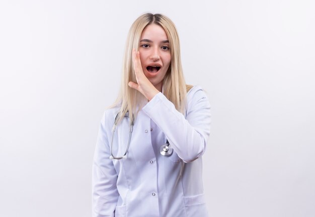 Doctor joven rubia con estetoscopio y bata médica en corsé dental susurra en la pared blanca aislada