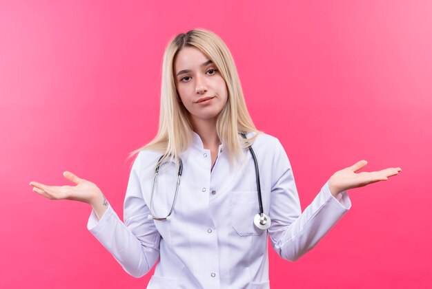 Doctor joven rubia con estetoscopio en bata médica extiende las manos sobre la pared rosa aislada