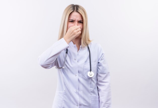Doctor joven rubia con estetoscopio y bata médica nariz cerrada en pared blanca aislada