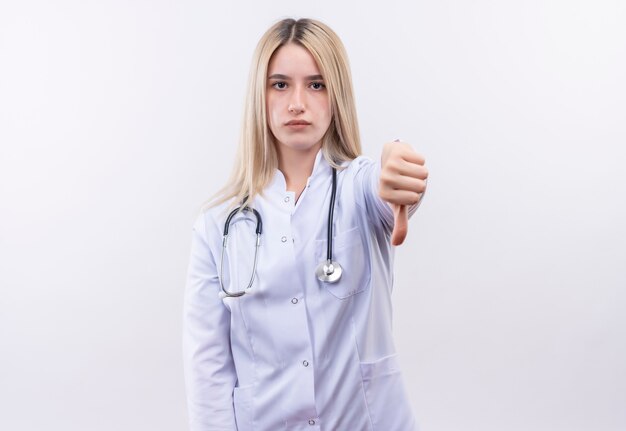 Doctor joven rubia con estetoscopio y bata médica con el pulgar hacia abajo en la pared blanca aislada