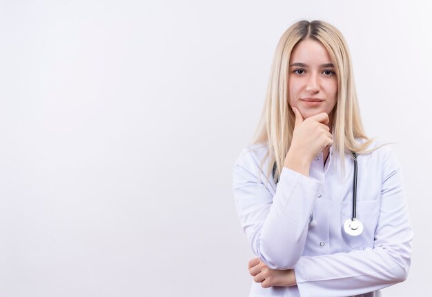 Doctor joven rubia con estetoscopio y bata médica puso su mano en la barbilla en la pared blanca aislada