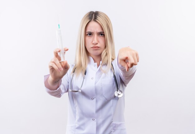 Doctor joven rubia con estetoscopio y bata médica sosteniendo una jeringa que le muestra un gesto en la pared blanca aislada