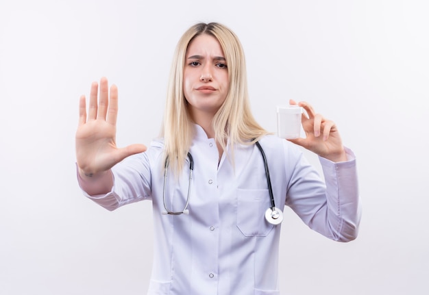 Doctor joven rubia con estetoscopio y bata médica sosteniendo lata vacía mostrando gesto de parada en la pared blanca aislada