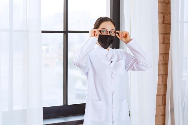 Doctor en máscara negra y anteojos de pie junto a la ventana.