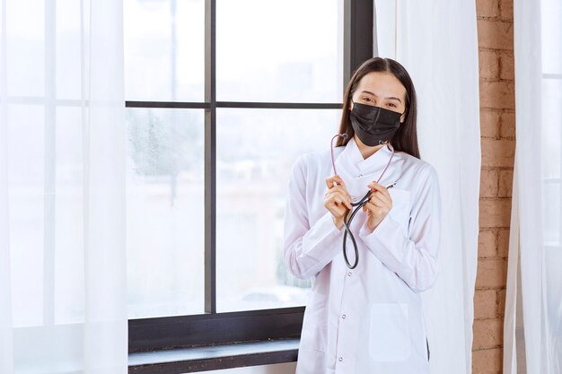 Doctor en máscara negra con un estetoscopio junto a la ventana.