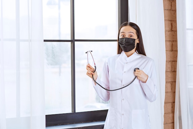 Doctor en máscara negra con un estetoscopio de pie junto a la ventana y controlando los latidos del corazón.