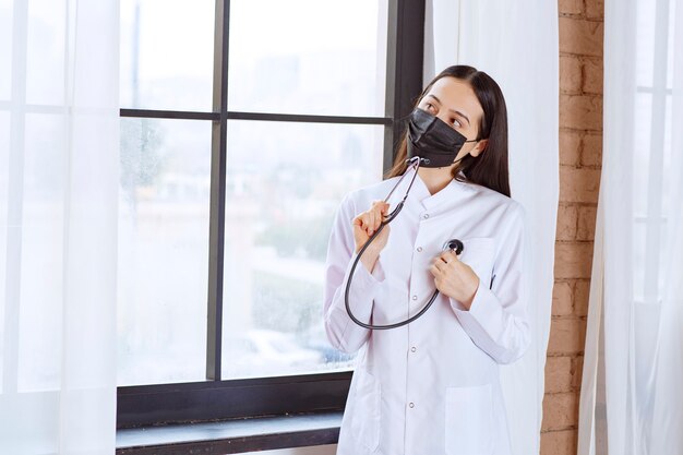 Doctor en máscara negra con un estetoscopio de pie junto a la ventana y controlando los latidos del corazón.