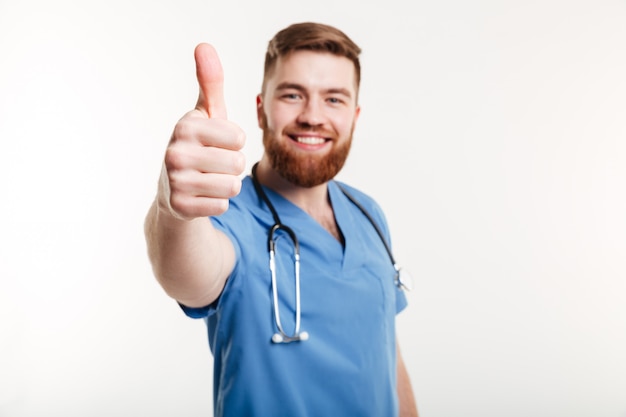 Doctor masculino alegre sonriente con el estetoscopio que muestra los pulgares para arriba