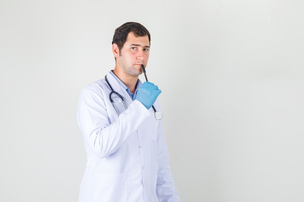Doctor masculino en bata blanca, guantes sosteniendo la pluma en los labios y mirando pensativo
