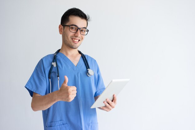 Doctor masculino feliz que sostiene la tableta y que muestra el pulgar para arriba