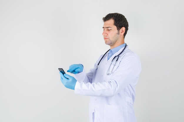 Doctor masculino tocando el teléfono inteligente con el dedo en bata blanca, guantes y mirando serio.
