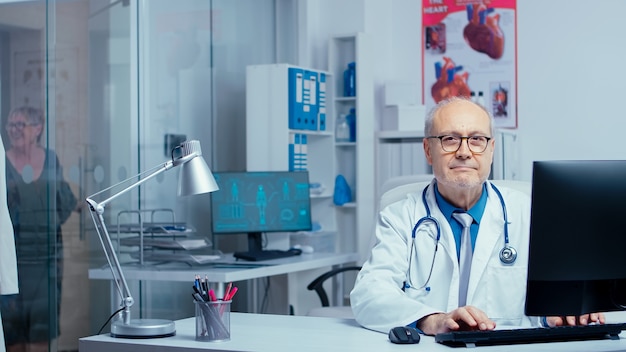 Doctor mirando a la cámara después de escribir en la computadora en una clínica de un hospital privado moderno, trabajando en la sala de consulta mientras la enfermera en la parte de atrás está hablando con un paciente. Paredes de vidrio