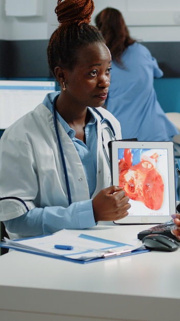 Doctor mostrando tableta con figura de corazón para chequeo cardiológico al paciente en consulta. Padre joven mirando un dispositivo con un órgano para la salud cardiovascular y del flujo sanguíneo.