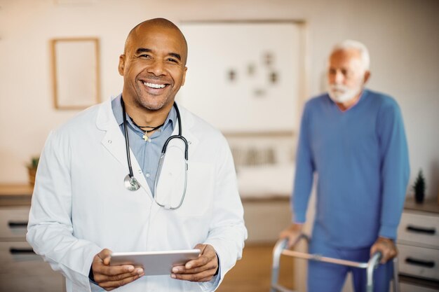 Doctor negro feliz que usa el panel táctil en el hogar de ancianos y mira la cámara