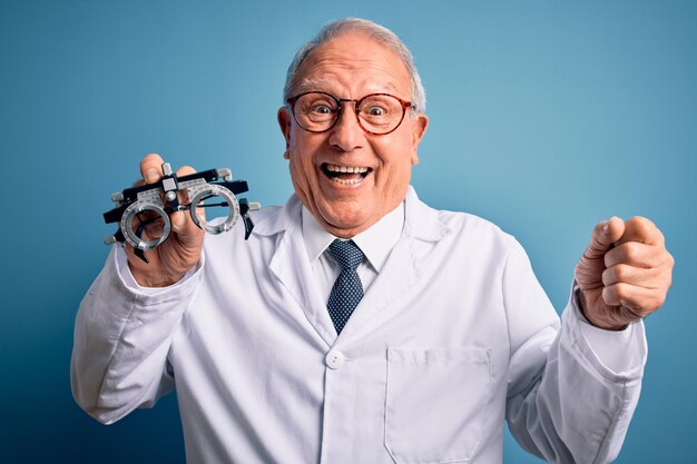 Doctor óptico de pelo gris senior hombre sosteniendo anteojos de optometrista sobre fondo azul gritando orgulloso y celebrando la victoria y el éxito muy emocionado animando la emoción