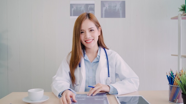 Doctor de sexo femenino asiático confiado que se sienta en el escritorio de oficina y que sonríe en la cámara