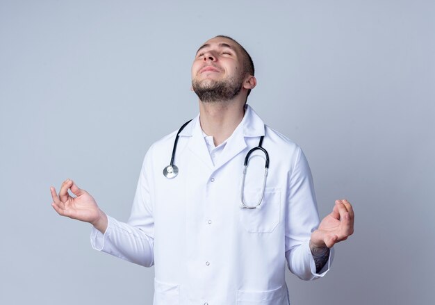 Doctor de sexo masculino joven complacido con bata médica y estetoscopio alrededor de su cuello meditando con los ojos cerrados aislados en la pared blanca