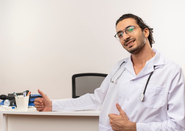 Doctor de sexo masculino joven complacido con gafas médicas vistiendo bata médica con estetoscopio de pie delante del escritorio