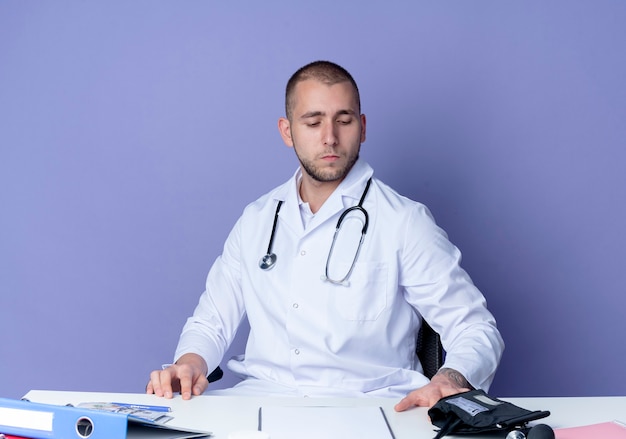 Doctor de sexo masculino joven que mira seriamente que lleva la bata médica y el estetoscopio sentado en el escritorio con herramientas de trabajo poniendo las manos sobre el escritorio y mirando hacia abajo en el escritorio aislado en la pared púrpura