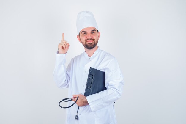 Doctor de sexo masculino joven que sostiene el portapapeles, el estetoscopio, apuntando hacia arriba con el uniforme blanco y mirando inteligente. vista frontal.