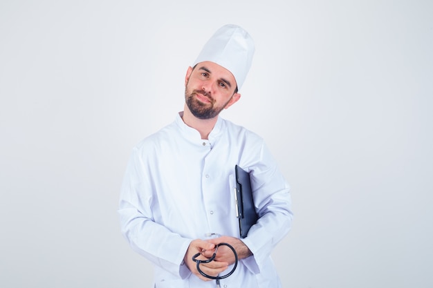 Doctor de sexo masculino joven que sostiene el portapapeles, estetoscopio en uniforme blanco y que parece suave. vista frontal.